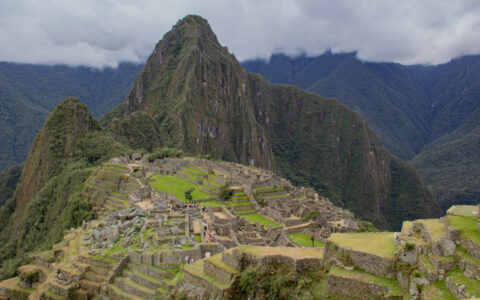 Looking down onto Machu Picchu citadel with tiered grass & stone levels high on a mountain plateau under a cloudy sky in Peru.