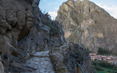 Mountainside cobblestone walkway in shadow, leading to view point of tall rocky mountain with beige carvings across the valley.