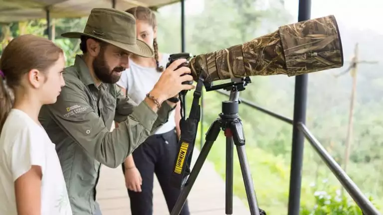 Man in green button-down shirt & hat adjusts camera with long camouflage lens sitting atop a tripod, as 2 girls look on.