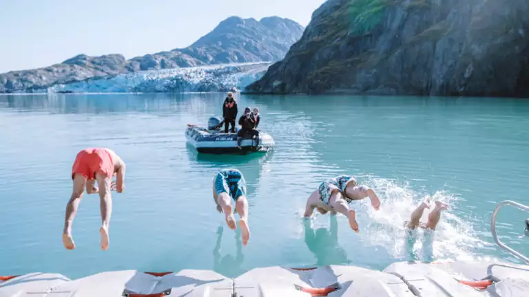 Guests leaping off the kayak towards zodiac platform into glacier bay for a polar plunge on a bright sunny day