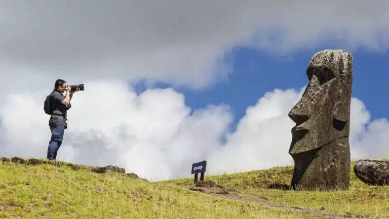 solo traveler takes a photograph of a rapa nui statue in easter island chile while standing on a grassy knoll on a partly sunny day