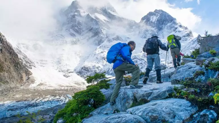 3 trekkers hike up white boulders on a sunny day with massive peaks in the background during the Torres del Paine Trek.