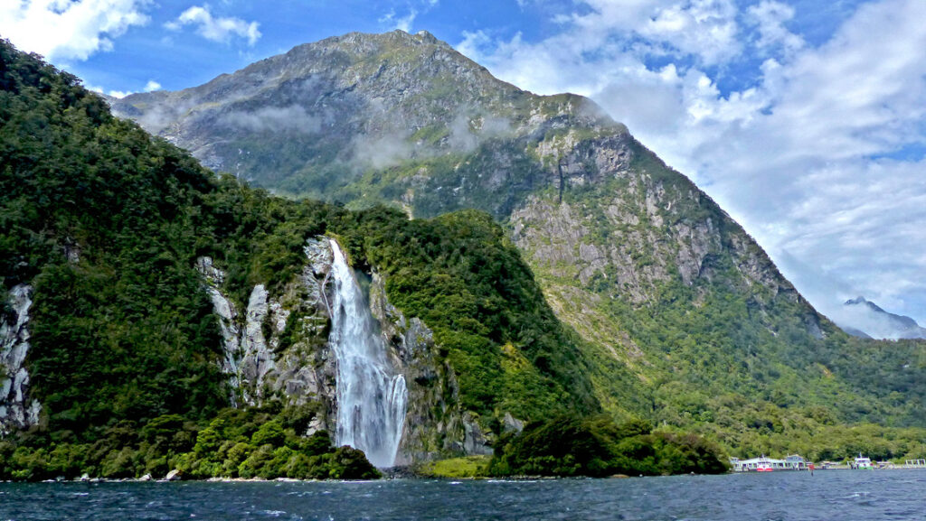 Waterfall cascades over ;ush green cliffs backed by a higher peak on a partly cloudy day in New Zealand.