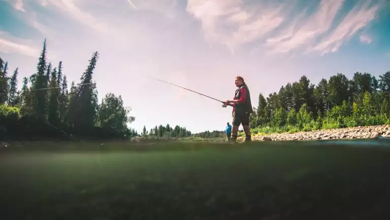 Woman stands at river's edge fishing while nearby man watches on a sunny day of the Within the Wild Alaska tour.