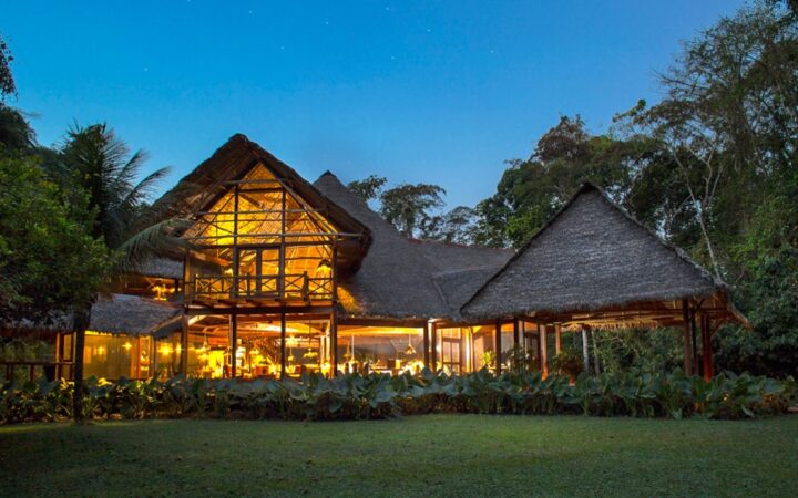 The exterior of the Inkaterra Reserva Amazonica at night, with thatched roofs and lights. It is an eco-luxury lodge in the heart of the Peruvian Amazon Rainforest, adjacent to the Tambopata National Park