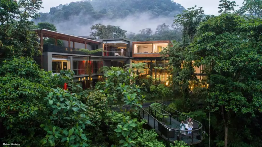 Exterior of Mashpi Lodge at dusk, with big glass windows surrounded by green vegetation & a viewing platform with 2 guests looking into the wilderness.