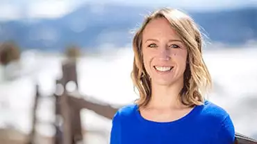 adventuresmith explorations adventure coordinator kelly gorrell wearing a blue shirt stands smiling on a sunny day in truckee with snow and mountains behind her