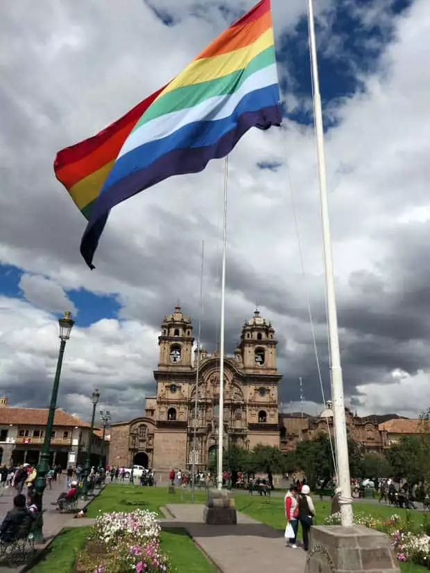 Peruvian flag flying in the middle of a courtyard with a colonial church in the background with many people meandering on a land tour in Peru.