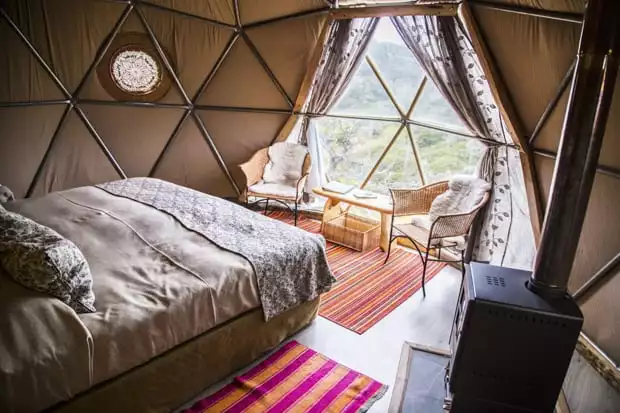 Inside of the dome EcoCamp with a bed, chairs, table and wood burning stove looking out into the forest of Torres del Paine.