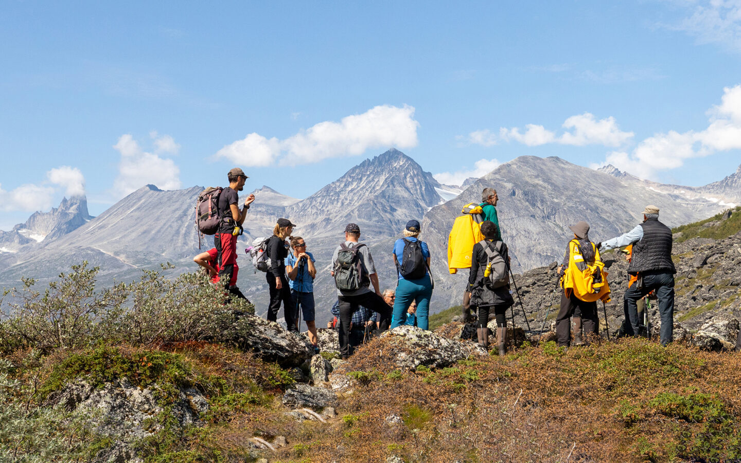 A group of adventure travelers stand high atop mountain range in Greenland looking at landscape that surrounds them.
