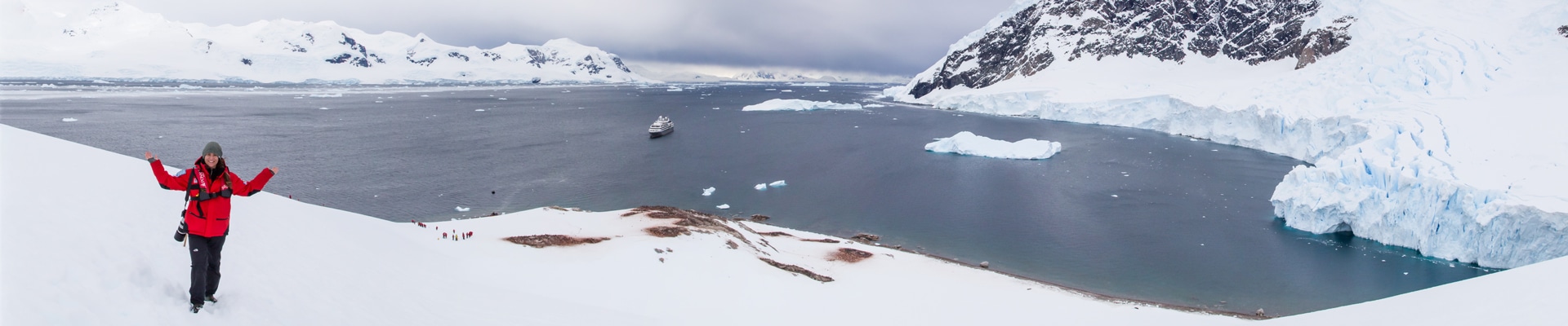 A female traveler in a red jacket stands on snow above a bay in Antarctica