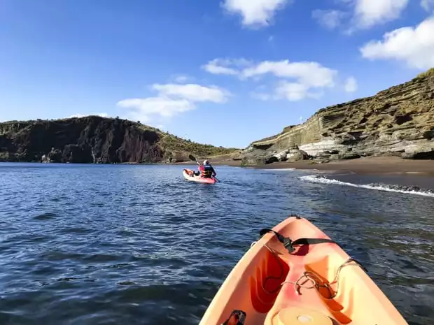 2 kayakers gliding off the shore of a sandy beach with bluffs.