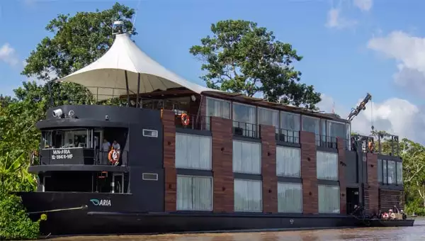 Aria luxury Amazon riverboat with dark steel and wooden exterior with large windows across three decks, cruising in the Peruvian Amazon on a sunny day.