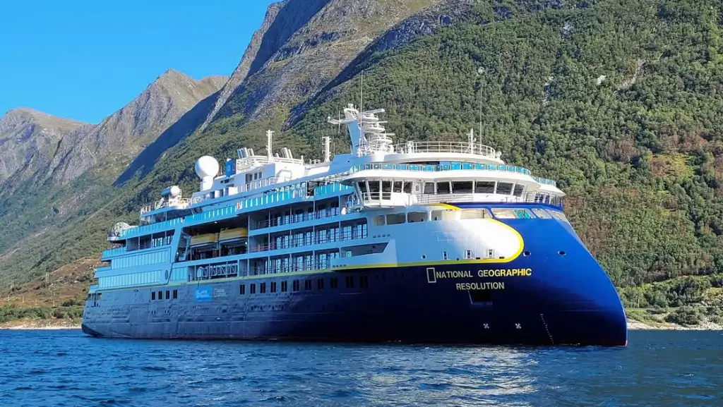 Large blue & white expedition ship Nat Geo Resolution with pointed bow sits in calm water beside green mountains on a sunny day.