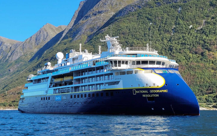 Large blue & white expedition ship Nat Geo Resolution with pointed bow sits in calm water beside green mountains on a sunny day.