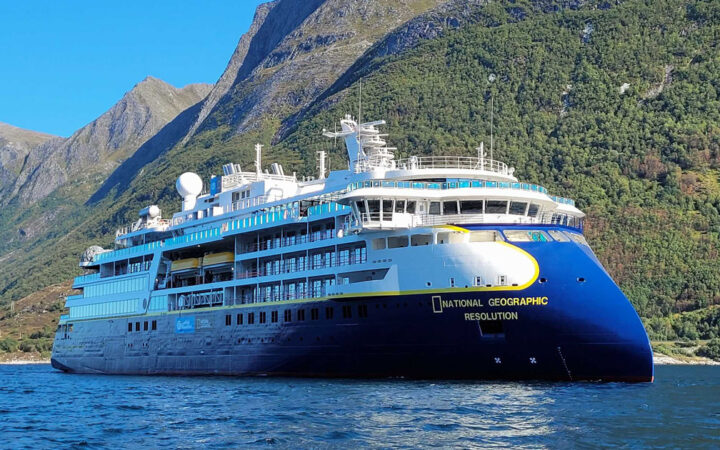 Large blue & white expedition ship Nat Geo Resolution with pointed bow sits in calm water beside green mountains on a sunny day.