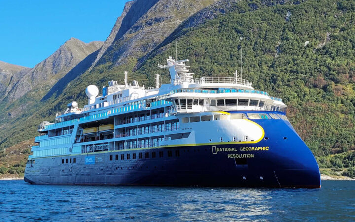 Large blue & white expedition ship Nat Geo Resolution with pointed bow sits in calm water beside green mountains on a sunny day.