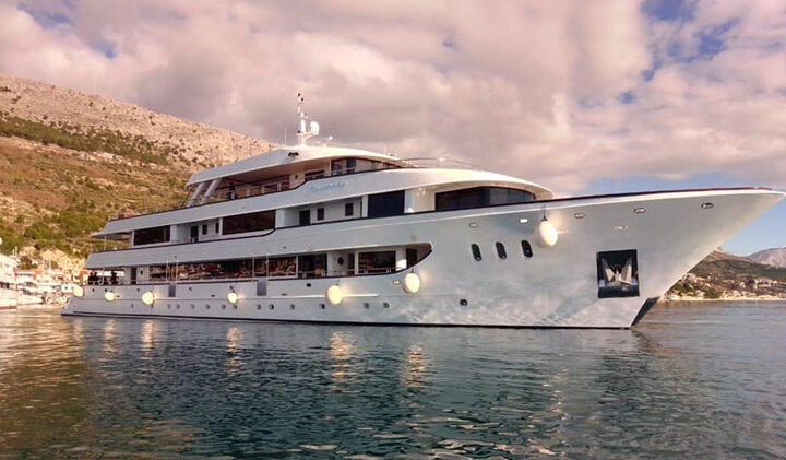 View from the water of the large white yacht Aurelia sitting in the harbor beside green hills on a partly cloudy day.