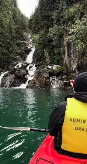 Sitting in a red double kayak you can see a kayaker wearing a yellow life jacket holding a camera to take a photo of a cliff side waterfall in front of him.