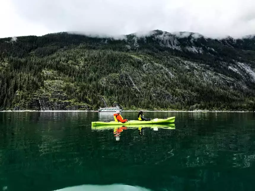 two paddlers in a tandem kayak floating on deep green water in front of mountain range and admiralty dream alaska small ship