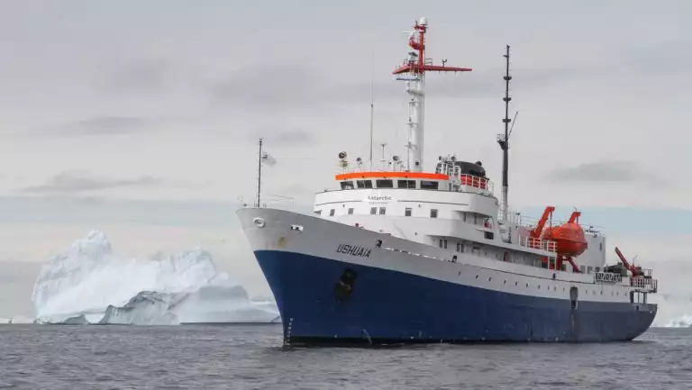Ushuaia small ship in blue & white sits beside a large iceberg on the Classic South Georgia small ship cruise in Antarctica.