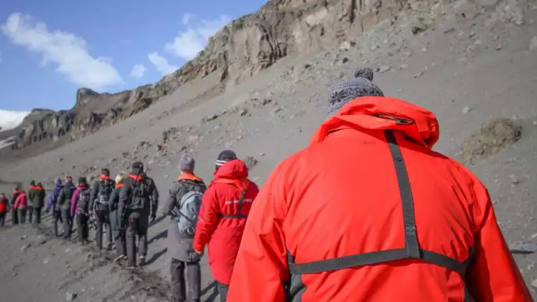 Line of Classic South Georgia cruise guests in colorful jackets walk a trail over gray dirt under blue skies.