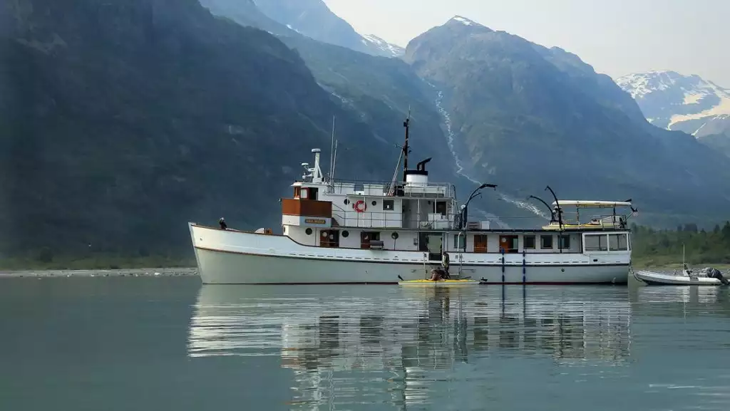 Alaska Small ship the Sea Wolf floats in Glacier Bay in front of a mountain range, kayakers paddle around the boat.