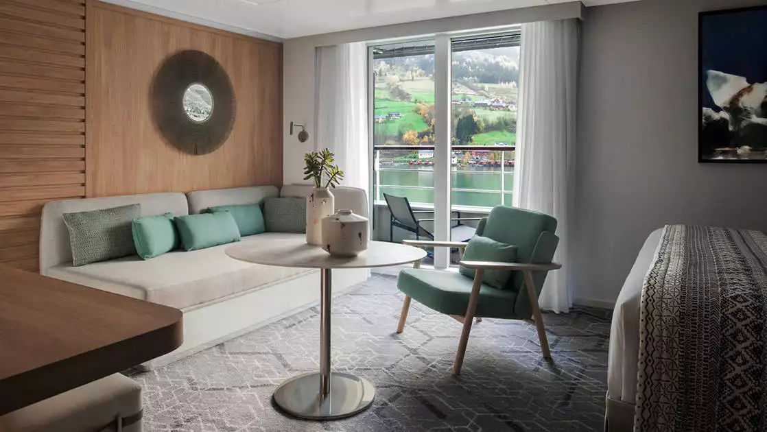 Privilege Suite aboard French luxury ship Le Champlain, with king bed, couch, table, chair & private balcony, plus natural-looking decor.
