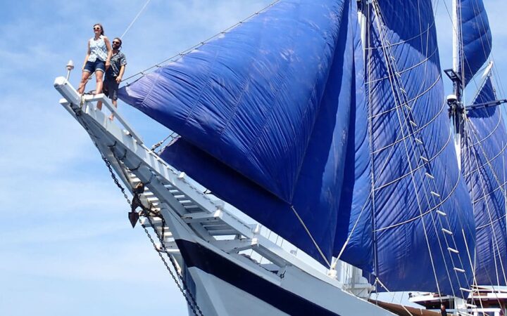 two people stand high in the air on the long wooden bow of the ombac putih indonesia small ship, its royal blue sails are up against the light blue sky