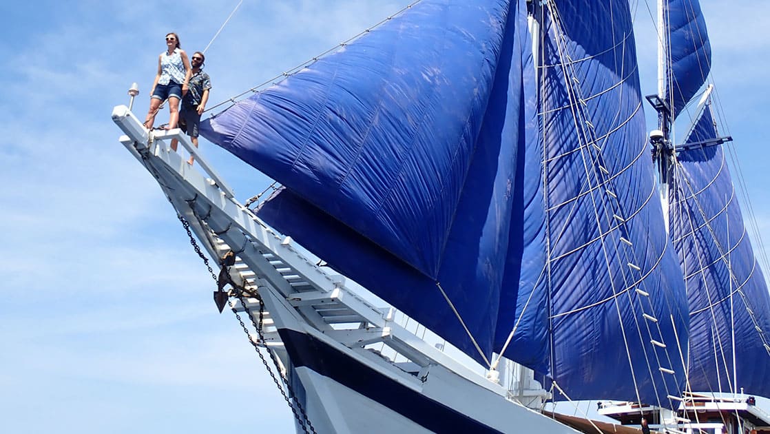 two people stand high in the air on the long wooden bow of the ombac putih indonesia small ship, its royal blue sails are up against the light blue sky