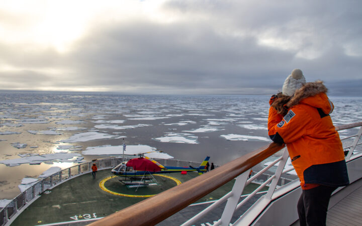 Woman in orange jacket & white knit hat stands on deck overlooking bow while cruising among icebergs on a cloudy day.