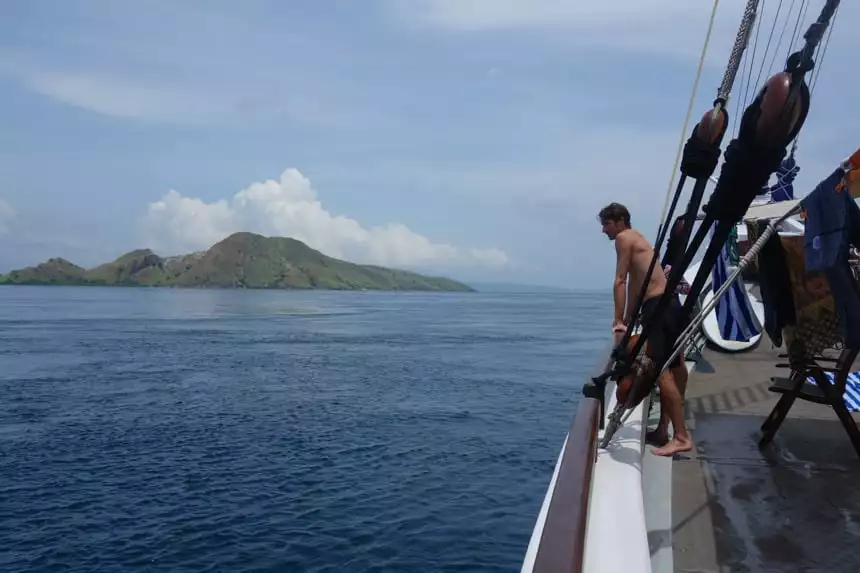 a cruise traveler leans on the ropes of the ombak putih's bow, looking at a snall green island in the distance of the blue ocean