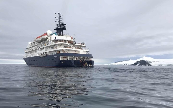 Luxury polar ship Hebridean Sky sits in calm, dark water under a cloudy sky, along a snow-covered shoreline in Antarctica, as guests embark a Zodiac at the stern.