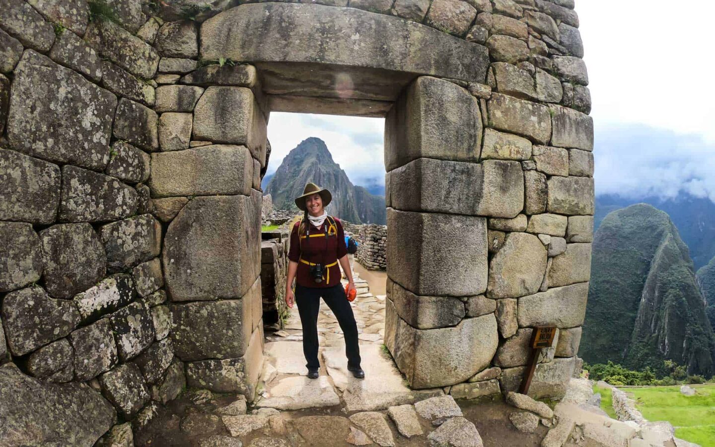 A female traveler stands inside a stone doorway of Machu Picchu Citadel, behind her through the door is a towering mountain peak.
