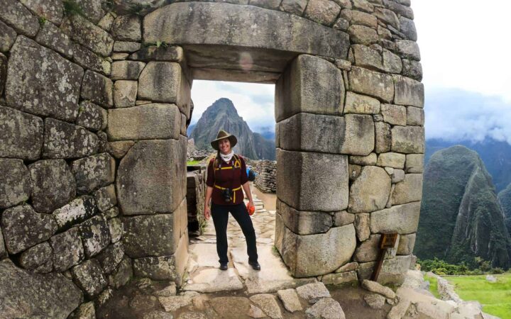 A female traveler stands inside a stone doorway of Machu Picchu Citadel, behind her through the door is a towering mountain peak.