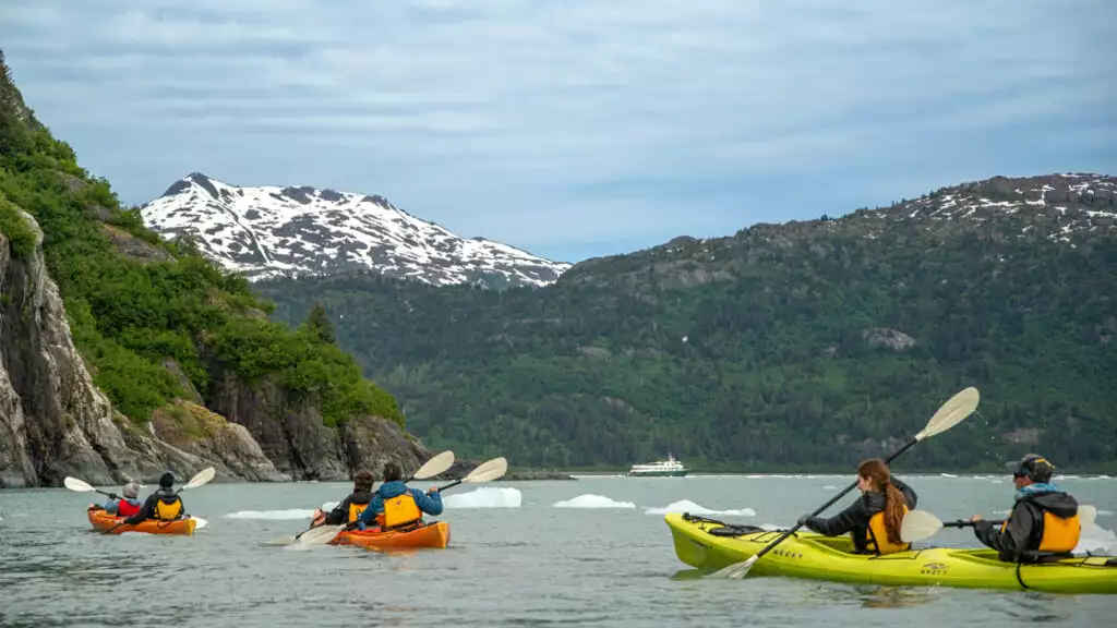 Kayakers in yellow lifevests, paddling through the bright tall mountains in alaska with snow off in the distance