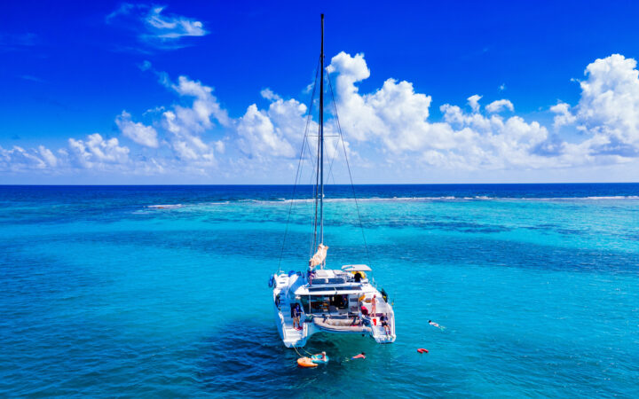 The exterior of a Belize charter ship, her blue sails are lowered on top of her all white deck as it floats in the teal ocean with guests swimming about