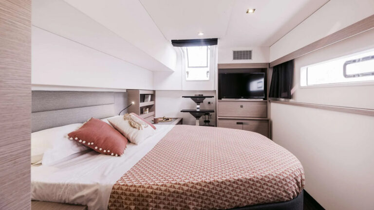 Cabin aboard The Big Leboatski with two smaller windows, a full bed, a tv, and multiple fans and storage spaces