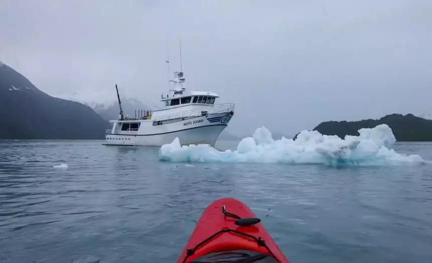 The tip of a red kayak floats in the foreground, infront of it floats an iceberg and the Misty Fjord ship on the islands whales glaciers cruise.