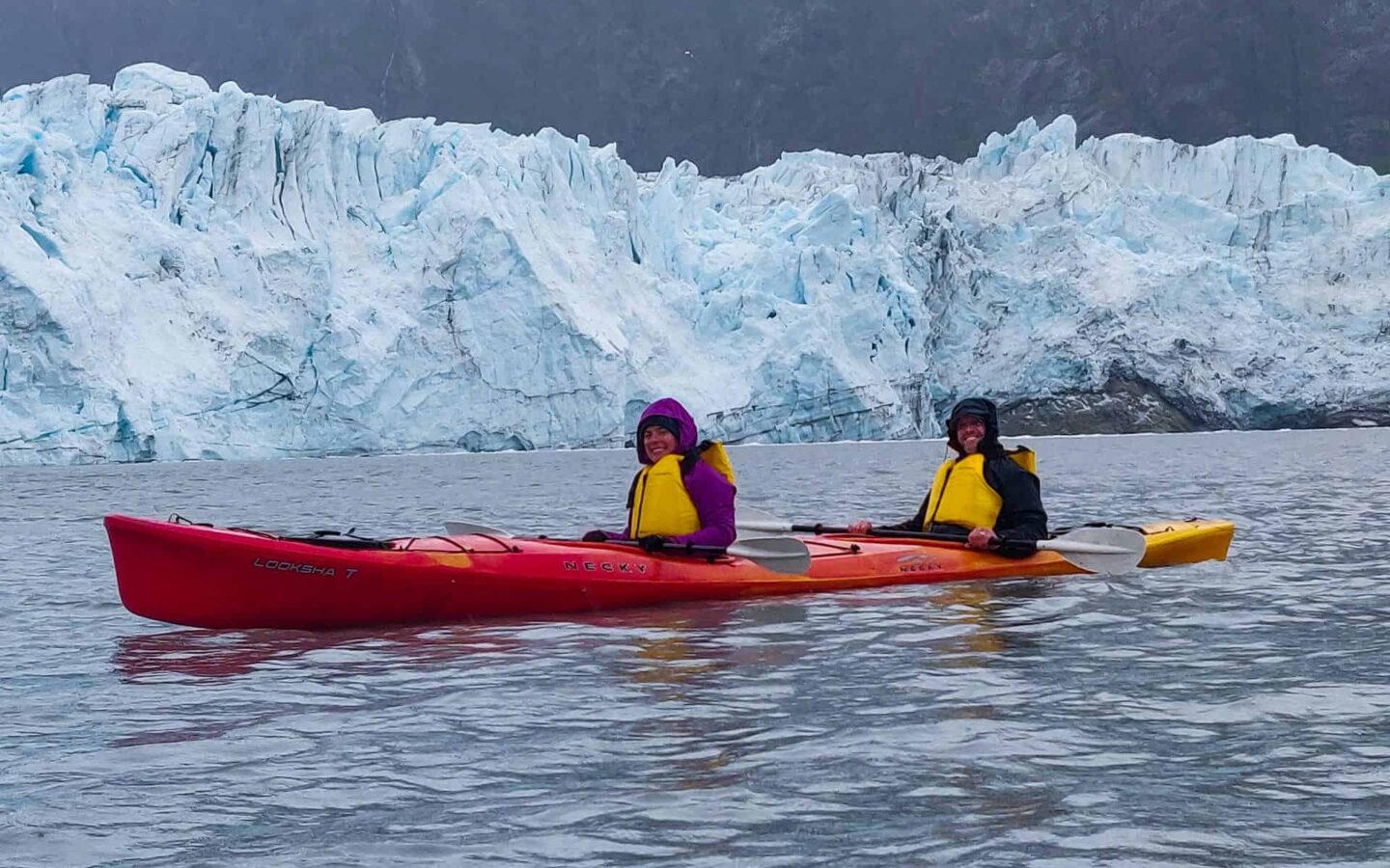 two happy guests paddle a red tandem kayak behind them is a jagged teal and white glacier, an activity done on slands whales and glacier small ship cruise