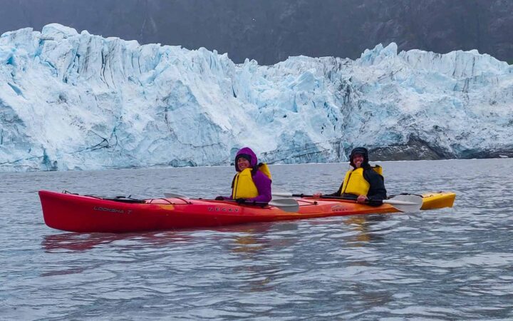 two happy guests paddle a red tandem kayak behind them is a jagged teal and white glacier, an activity done on slands whales and glacier small ship cruise