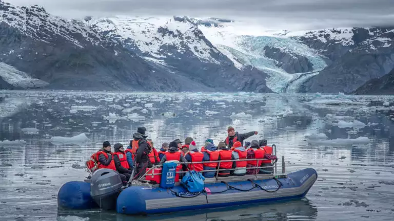 Explorers with red lifevests navigating through the icy still waters next to the run off of a large glacier off in the distance