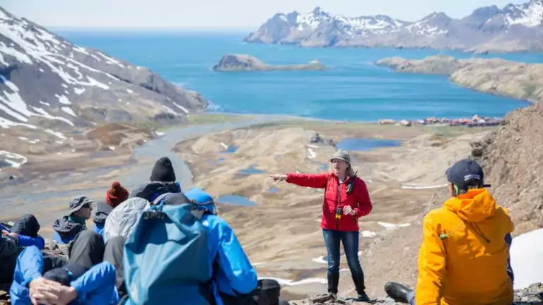 A woman in a red jacket stands & points in front of a group sitting on top of a tan mountain range during the South Georgia Antarctic Odyssey Cruise.