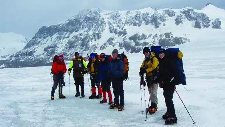 Group of polar travelers with large packs & trekking poles stand atop a snowfield during the Shackleton Crossing on South Georgia Island.