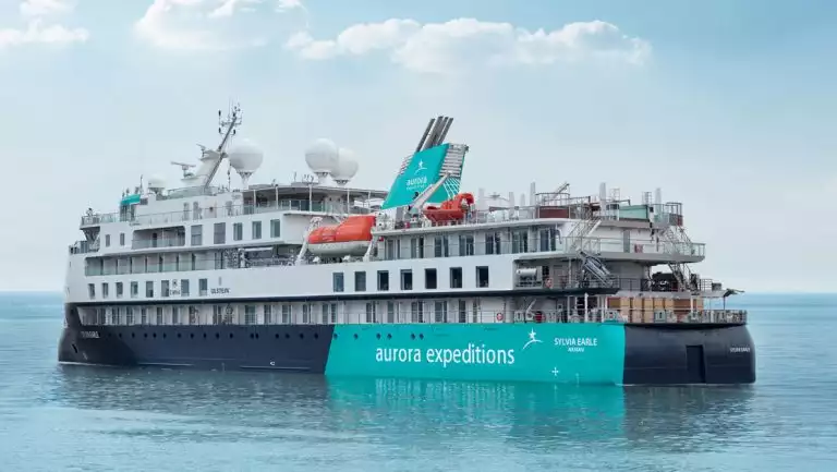 Sylvia Earle ship sitting in calm water, with blue & teal exterior with star logo, 5 white upper decks & 2 orange life boats.