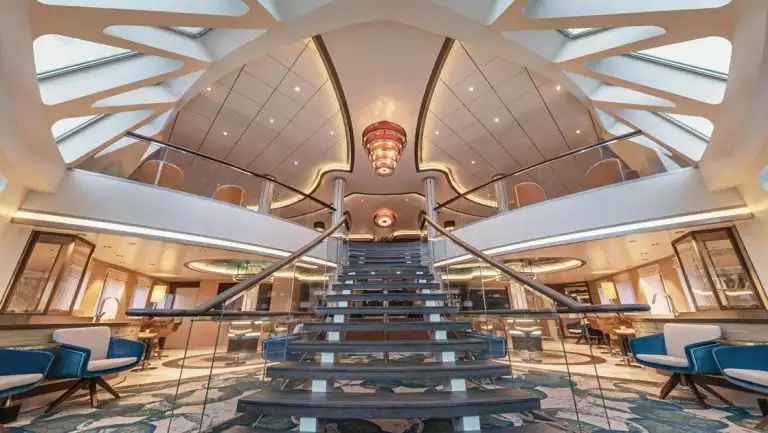 Library on Sylvia Earle ship with central staircase, blue & white carpet, glass rectangle ceiling tiles, chandelier & 2 floors.