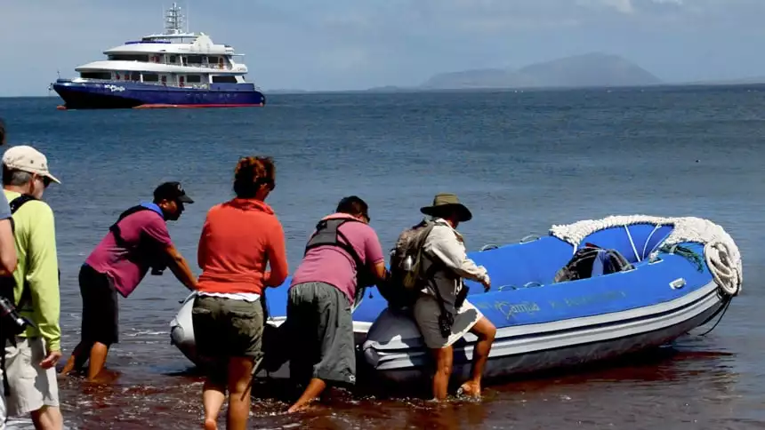 a group of travelers board an inflatable skiff or panga from the red sand beach on Rabida Island, in the distance the Camila galapagos ship floats in the ocean