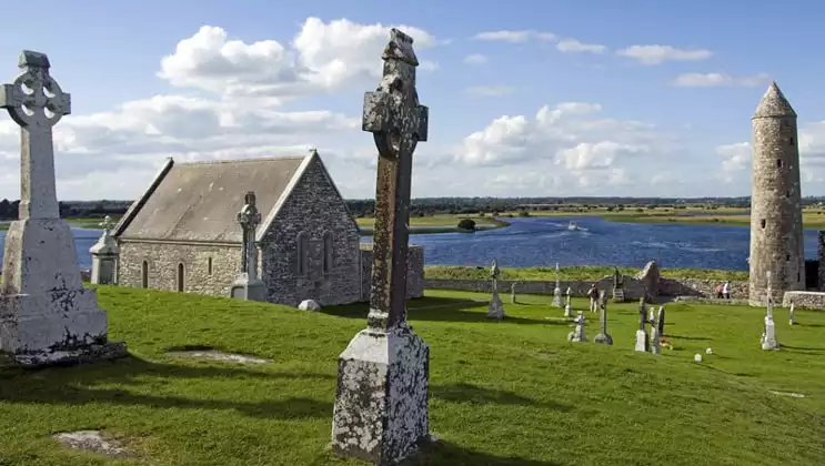 Small old stone church by green grass, stone crosses & calm river on a sunny day of the Classic Ireland River Cruise.