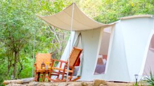 Canvas green & beige tent at Camp Cecil de la Sierra, with awning, 2 wooden rocking chairs & a table on a sunny day.