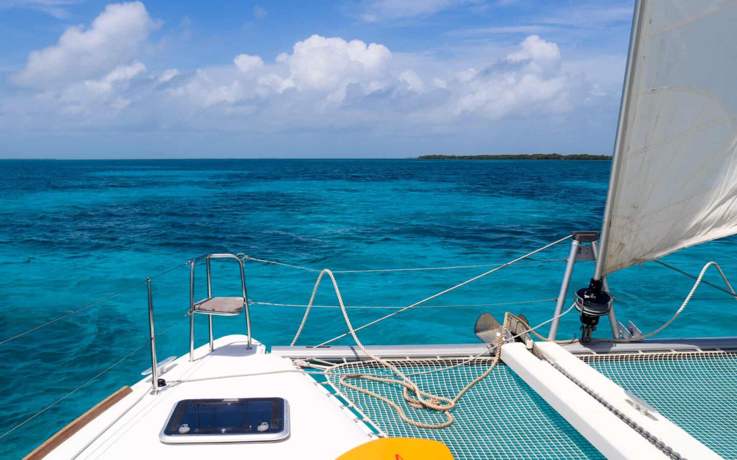the view from the rear of a Belize catamaran charter.The white netting and white sails sit against a crystal blue and teal ocean horizon and cloudy sky